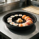 Plateau à sushi rond Ø 280 mm en polystyrène noir garni de sushis disposés en couronne, en cuisine japonaise
